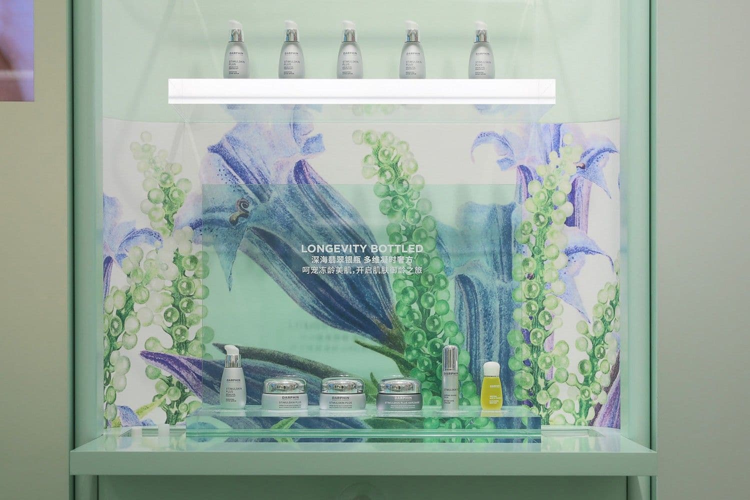 Glass display shelf lit from above, holding Darphin skincare bottles and jars in front of an illustrated underwater floral panel.