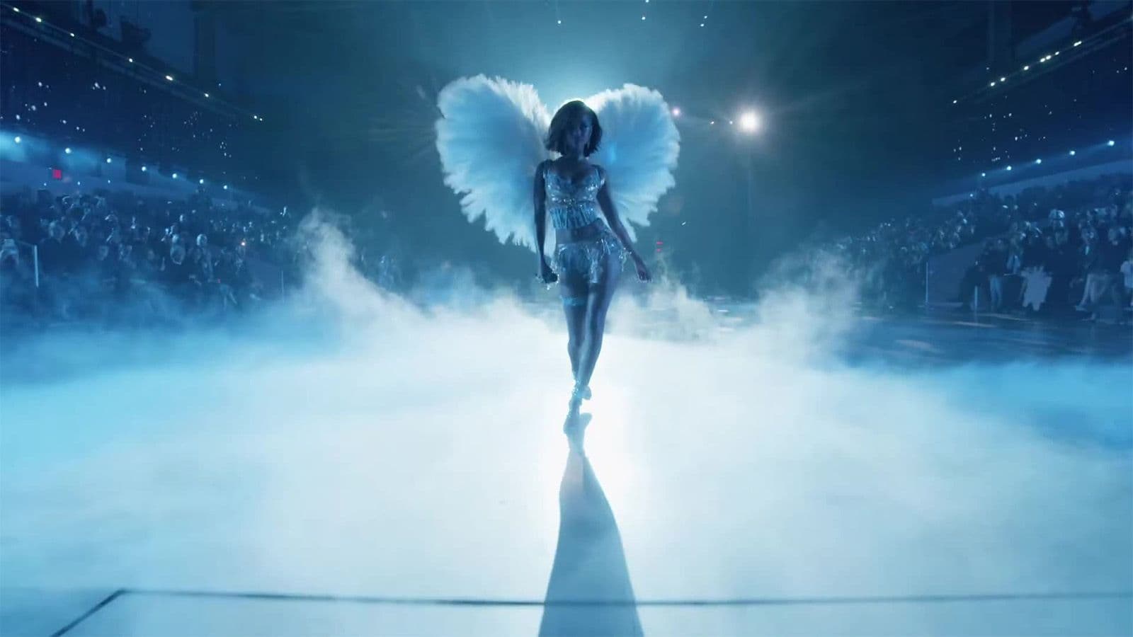 A model in a winged costume walks through fog toward the camera on a blue-lit runway packed with audience.