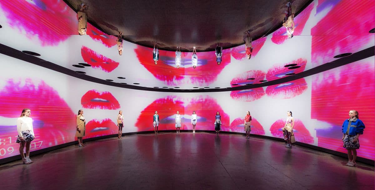 Models standing in a circular runway space with oversized pink lip graphics projected on curved screens overhead and around the walls.