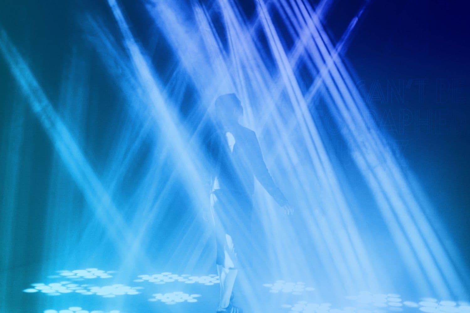 Dark room with sweeping blue light beams and a silhouetted figure walking through the installation.