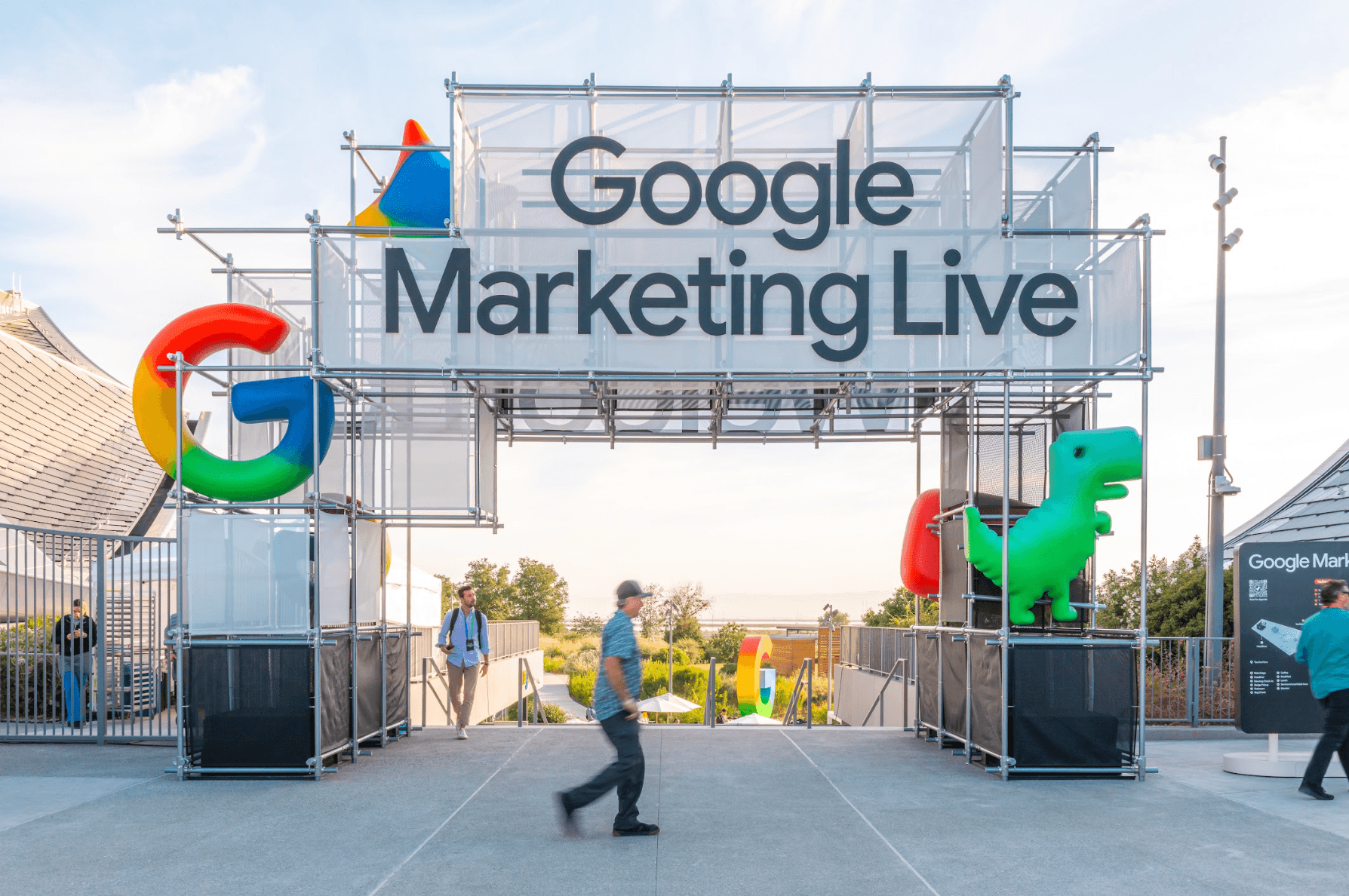 Entrance arch reading "Google Marketing Live" with a colorful Google G logo and an inflatable green dinosaur, attendees walking underneath.
