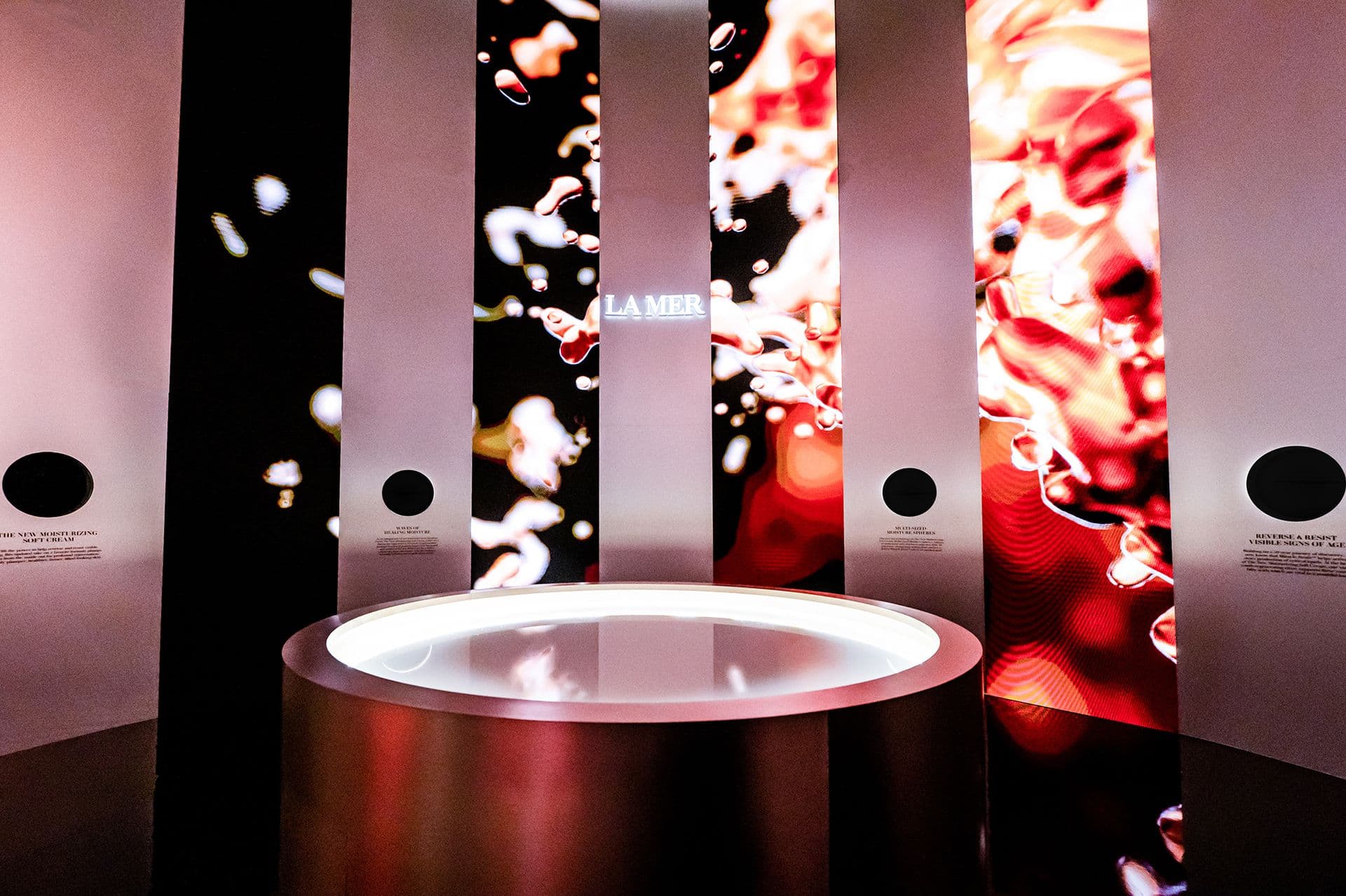 Vertical striped display columns in coral and red tones above a glowing circular table with La Mer branding.