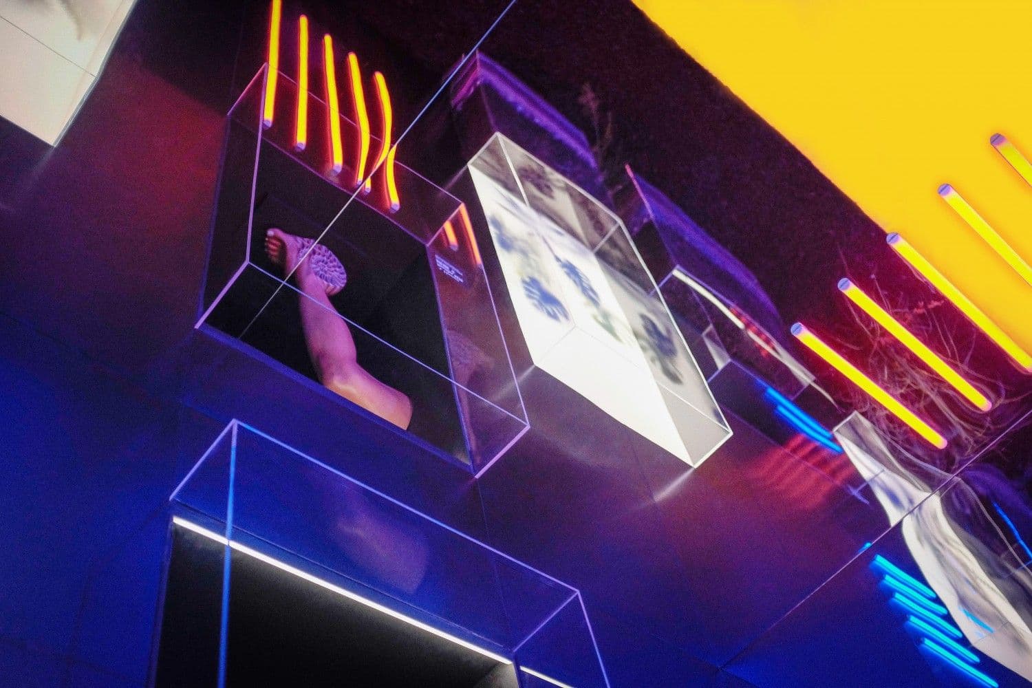 Low-angle view of glass display cubes and neon tubes in a purple and yellow lit space, with an arm reaching from one cube.