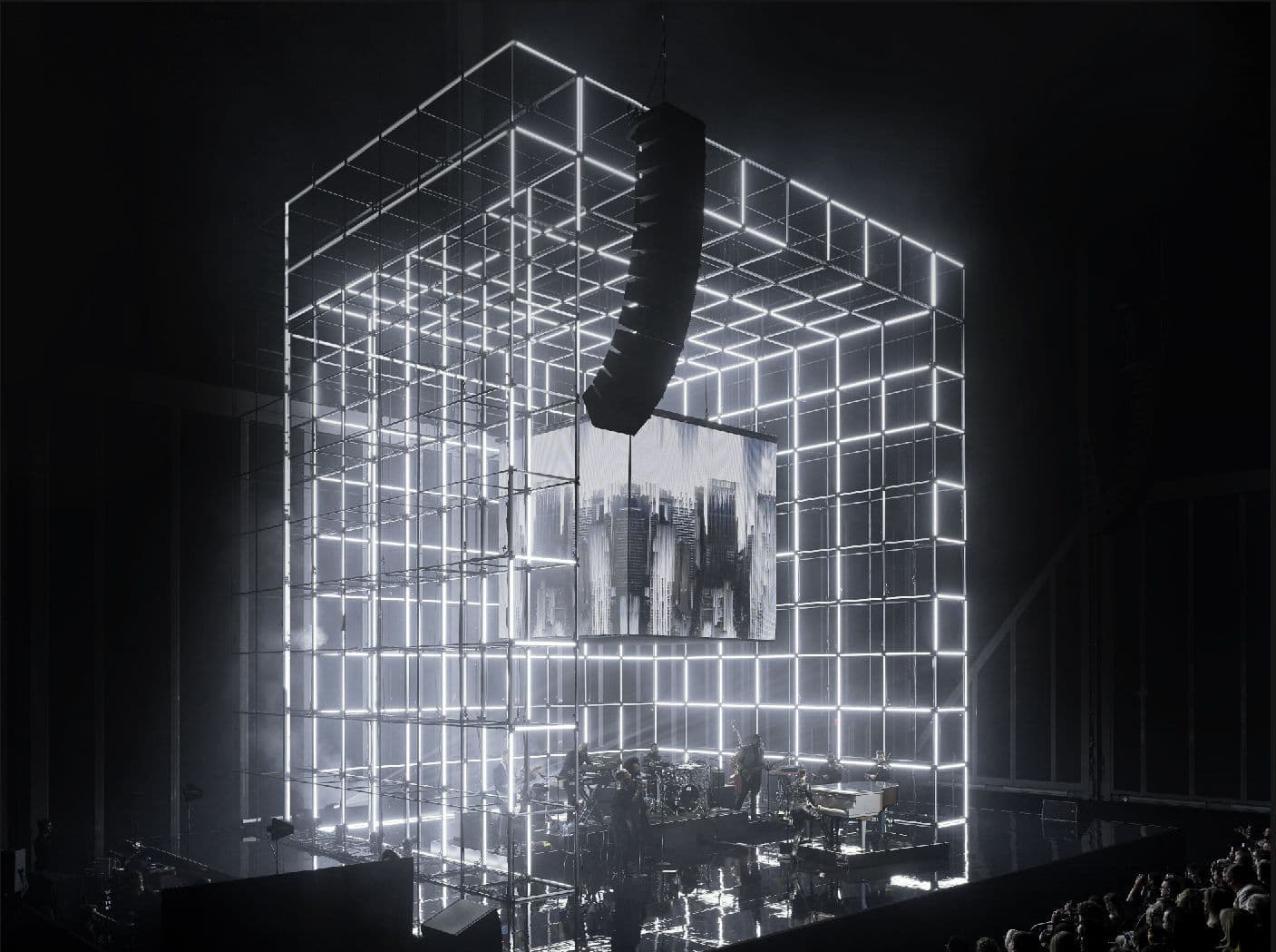 Wireframe cube stage structure with a dim video panel showing silhouetted figures at its center, rows of seated audience visible in the foreground.