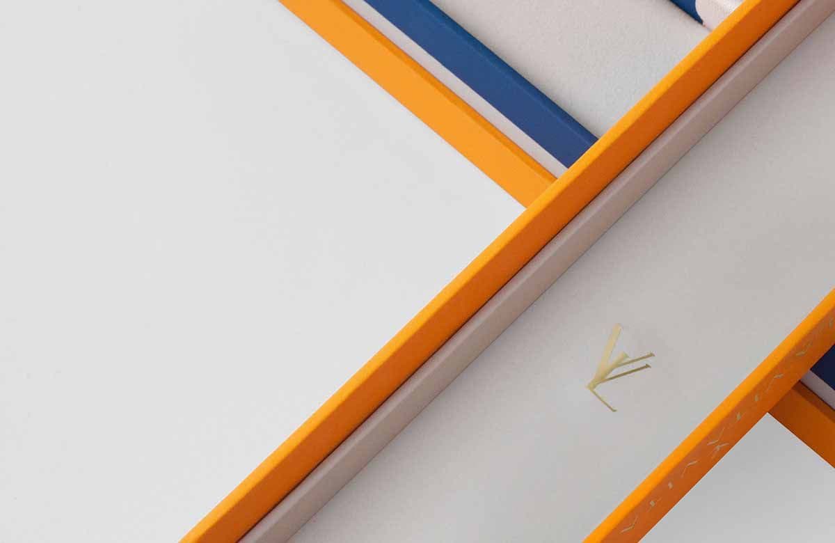 Diagonal close-up of orange and navy blue box edges meeting at a point, with a small gold logo mark on the gray panel between them.