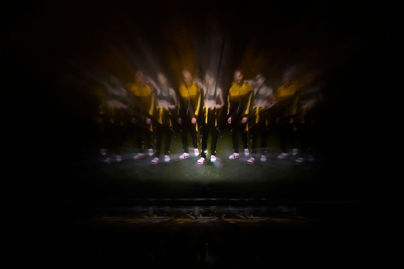 Silhouetted performers on an elevated stage engulfed in bright stage lighting, viewed from above a crowd at night.