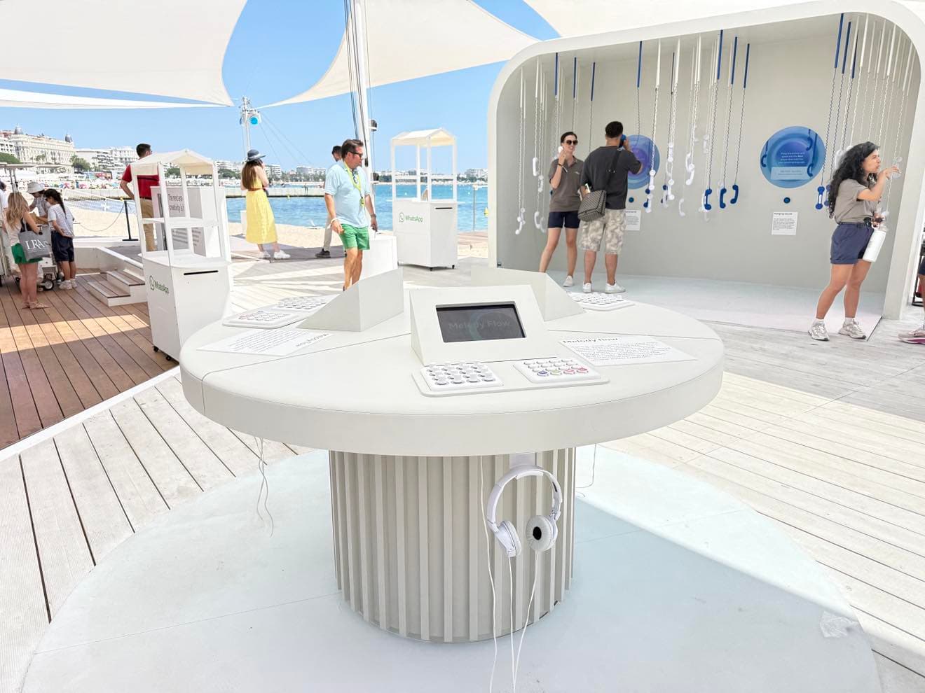 A round white interactive kiosk with a touchscreen and headphones on a beachside wooden deck, with a tented pavilion and visitors in the background.