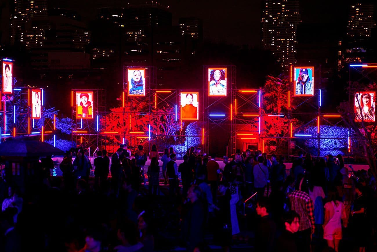 Crowd gathered at an outdoor Tommy Hilfiger event lit with red and blue neon amid illuminated city skyscrapers.