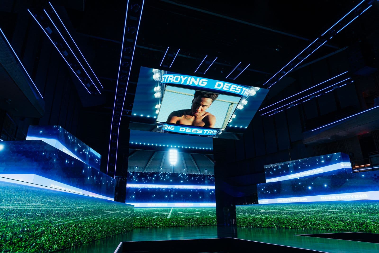 Wide shot of an arena stage with blue LED screens displaying a stadium scene above the crowd.