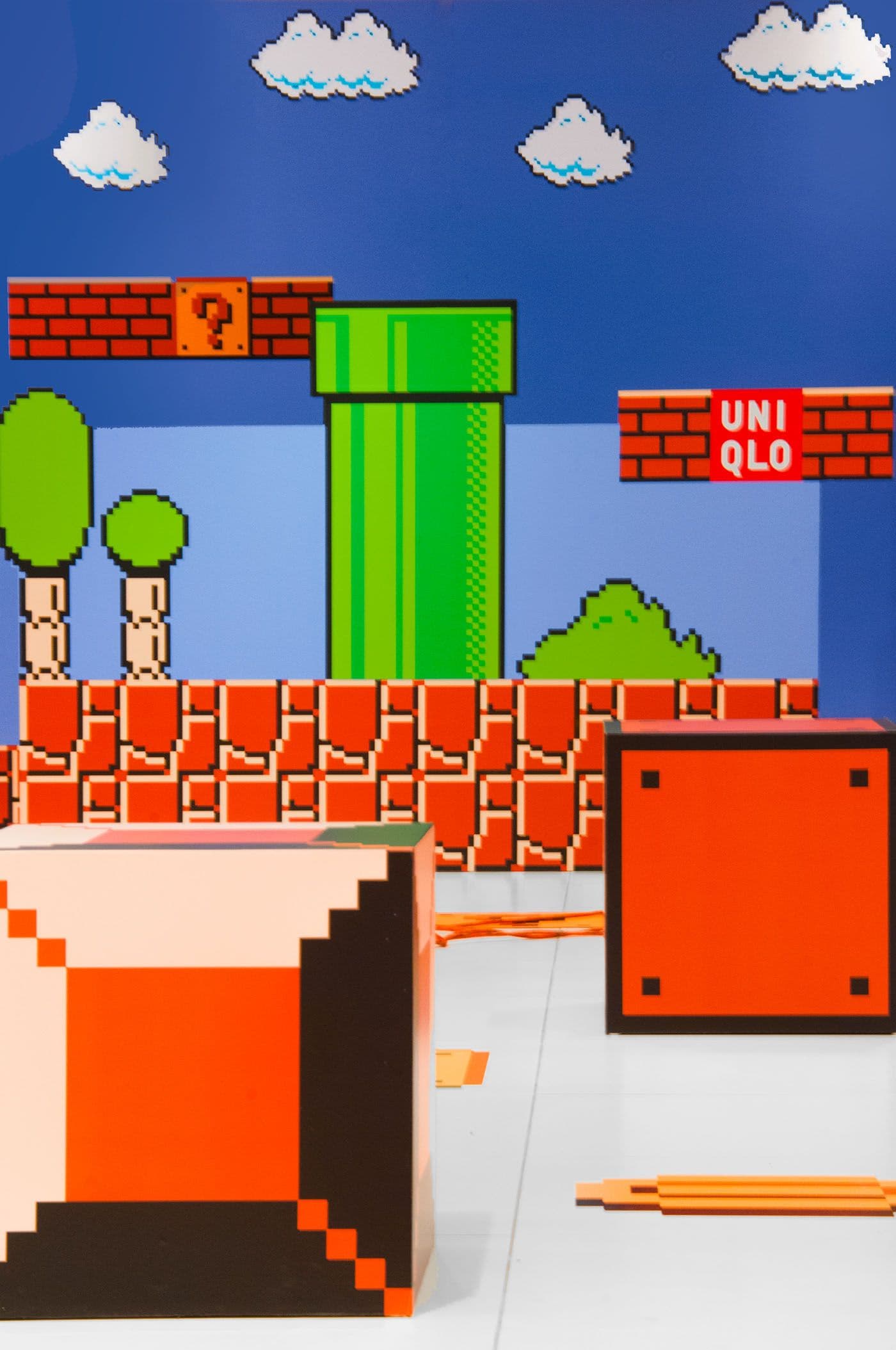 Pixel-art Super Mario Bros scene mural with brick blocks, a green pipe, clouds and trees, a red UNIQLO sign, and orange block sculptures in front.