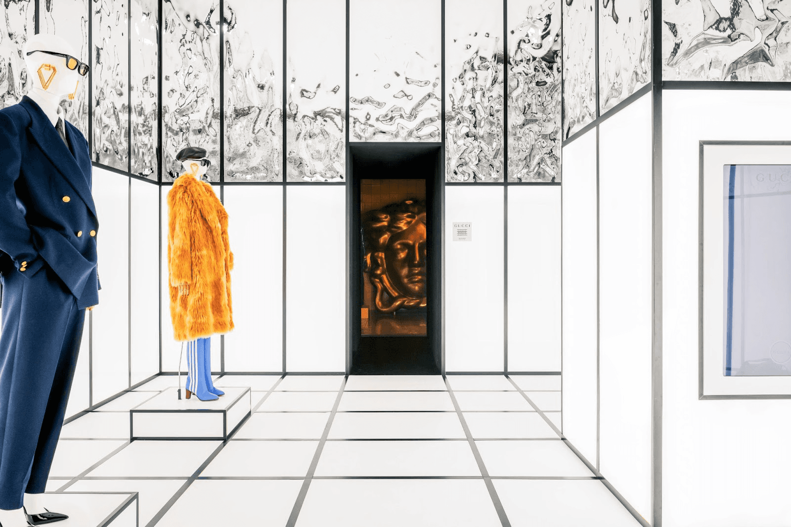 Two mannequins, in a navy suit and a mustard fur coat, standing in a mirrored checkerboard room with a glowing face visible through a doorway.