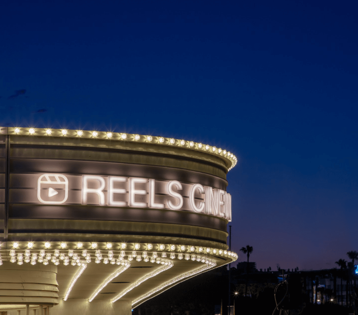 A round marquee-style theater sign lit up at night reading 'Reels Cinema' against a dark blue sky.