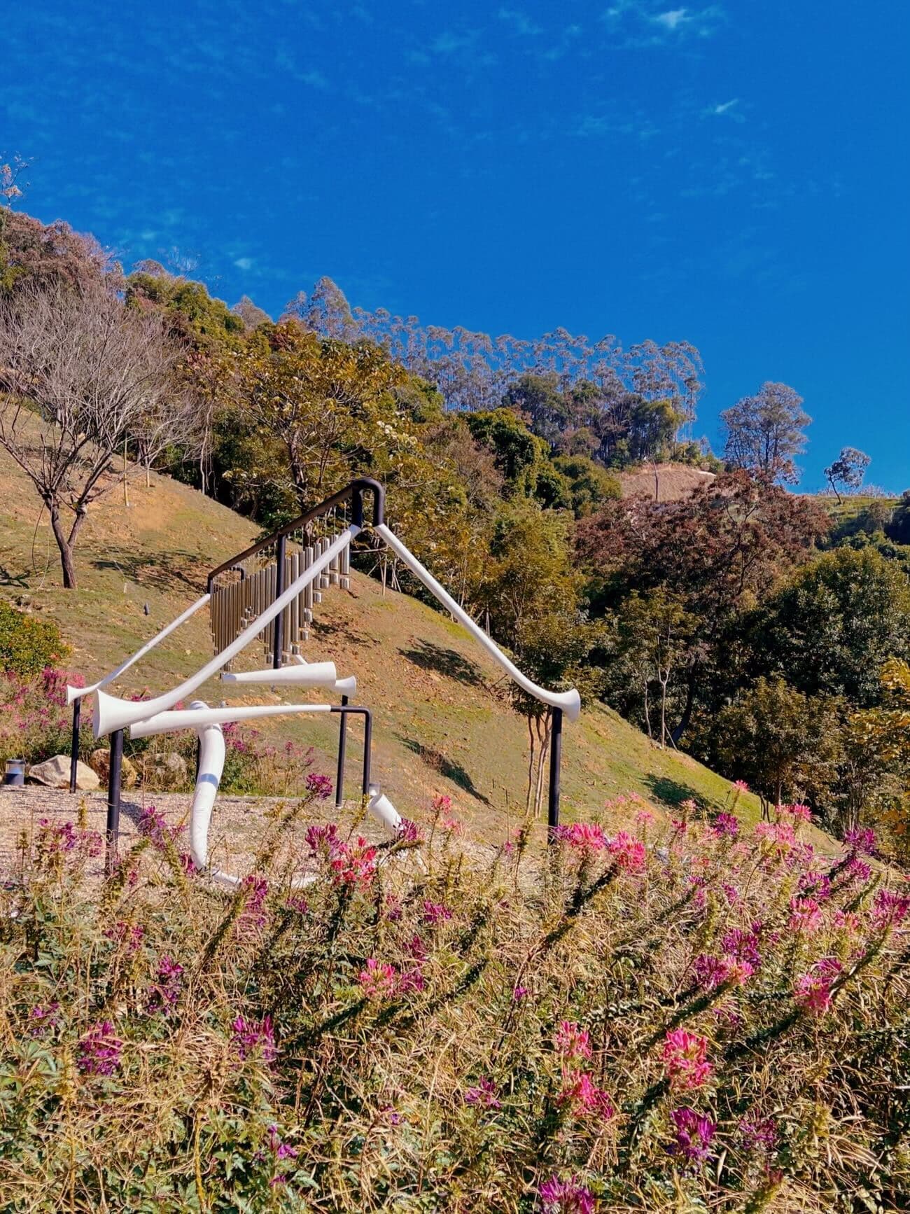 White tubular slide and climbing structure on a grassy hillside with flowering pink shrubs in the foreground.