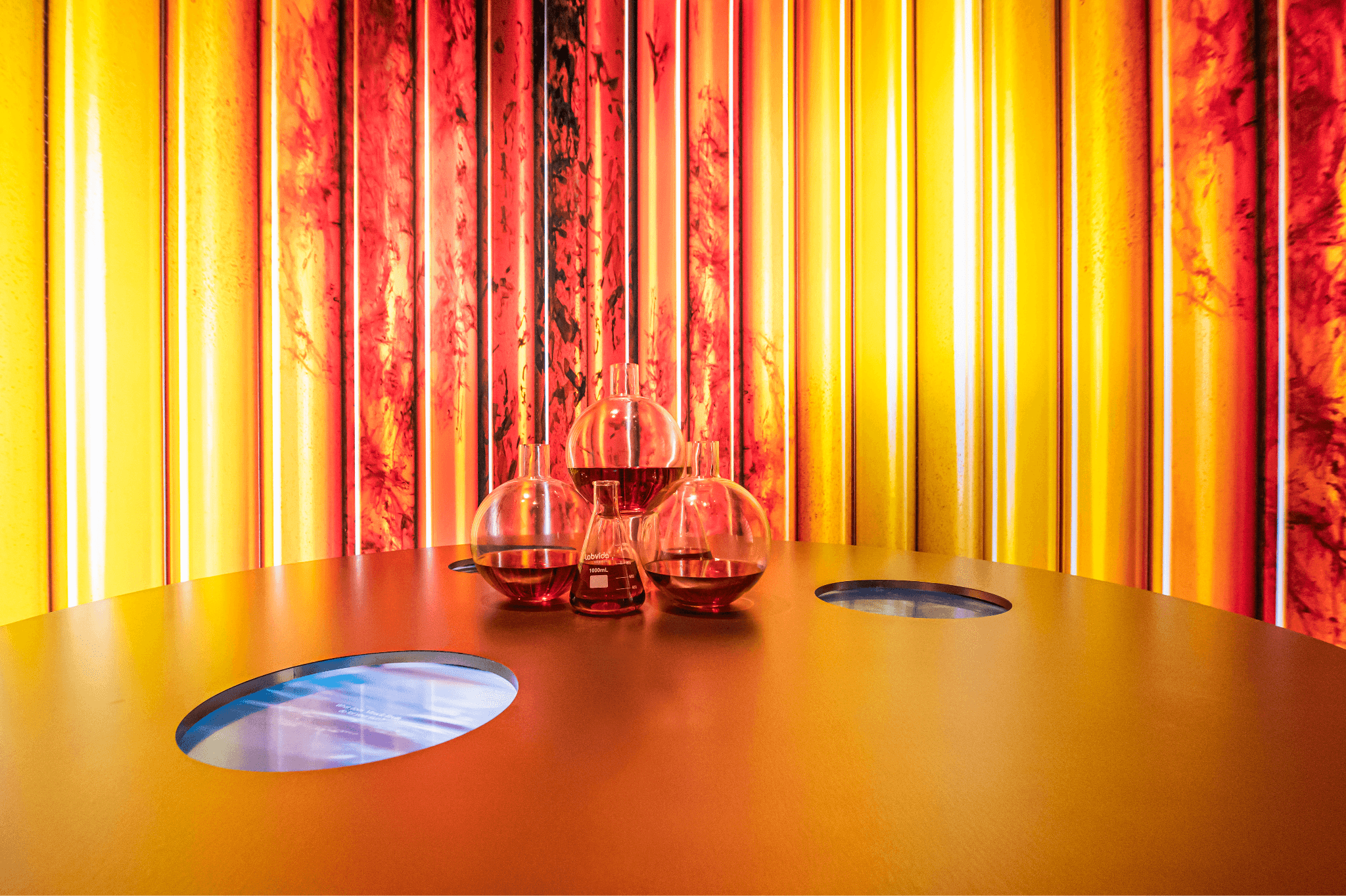 Wide view of red and orange striped display columns around a circular table holding glass vessels, with illuminated floor discs.