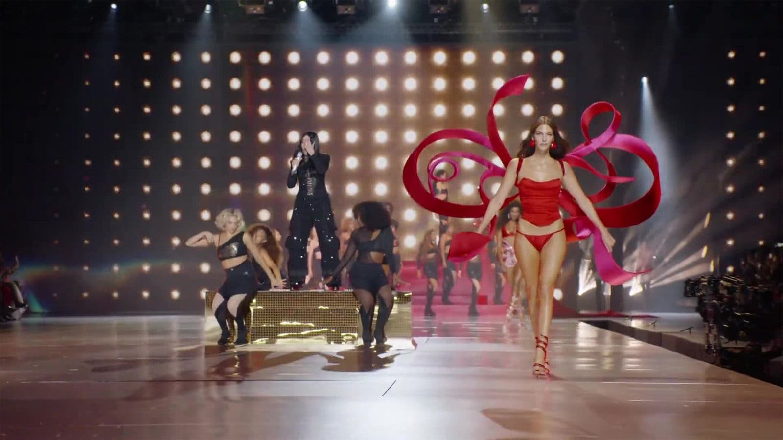 A model in a red gown trails a long red ribbon down the runway past dancers seated on a bench under stage lights.