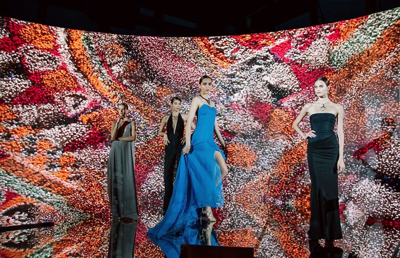 Three models in black, red, and blue gowns standing on a runway in front of a curved LED screen showing an abstract red and orange pattern.