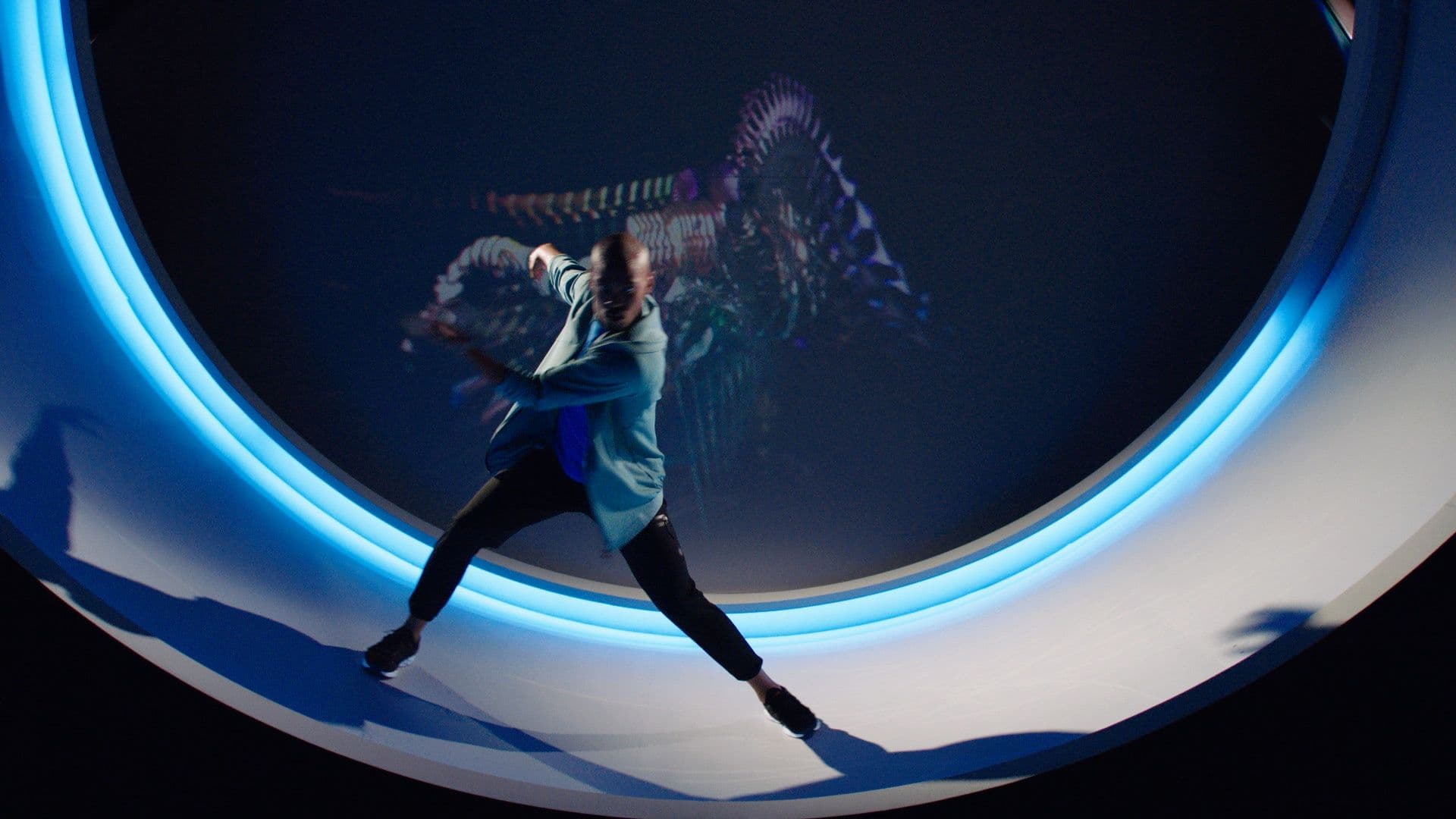 Dancer captured mid-motion inside a blue-lit circular tunnel set, arm raised with a motion-blurred silhouette overhead.
