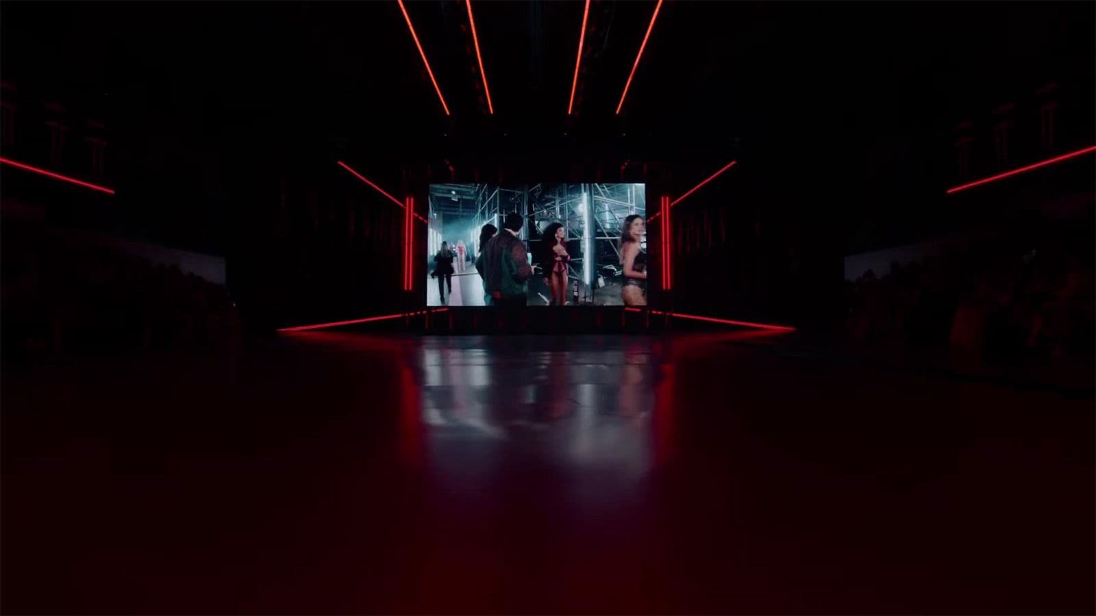 A large video screen displaying dancers glows at the end of a dark runway framed by red laser light beams.