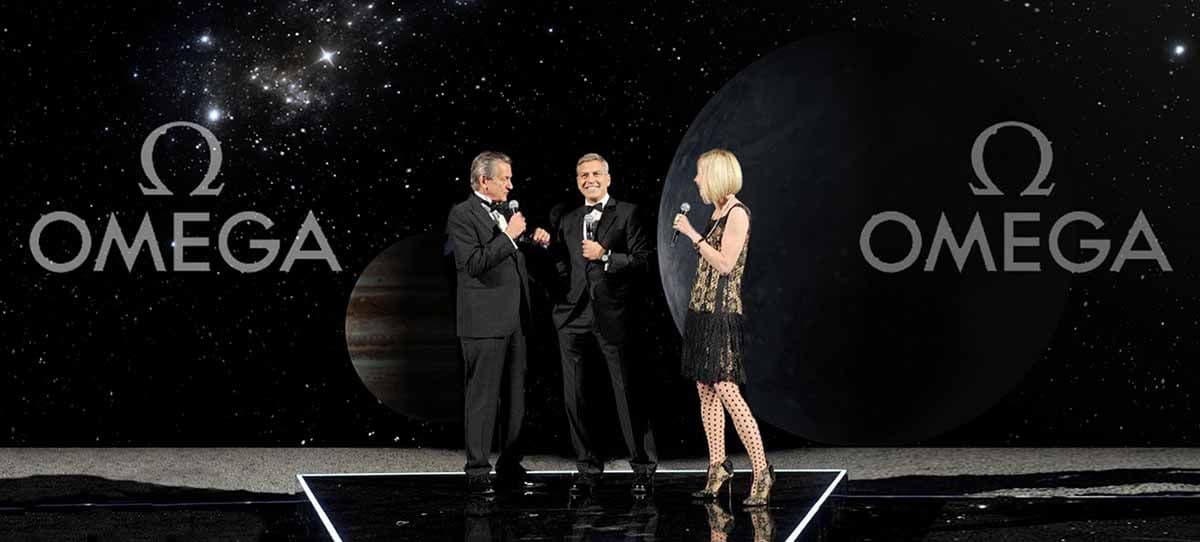 Three people standing on a stage in front of two large OMEGA logos against a starfield backdrop.