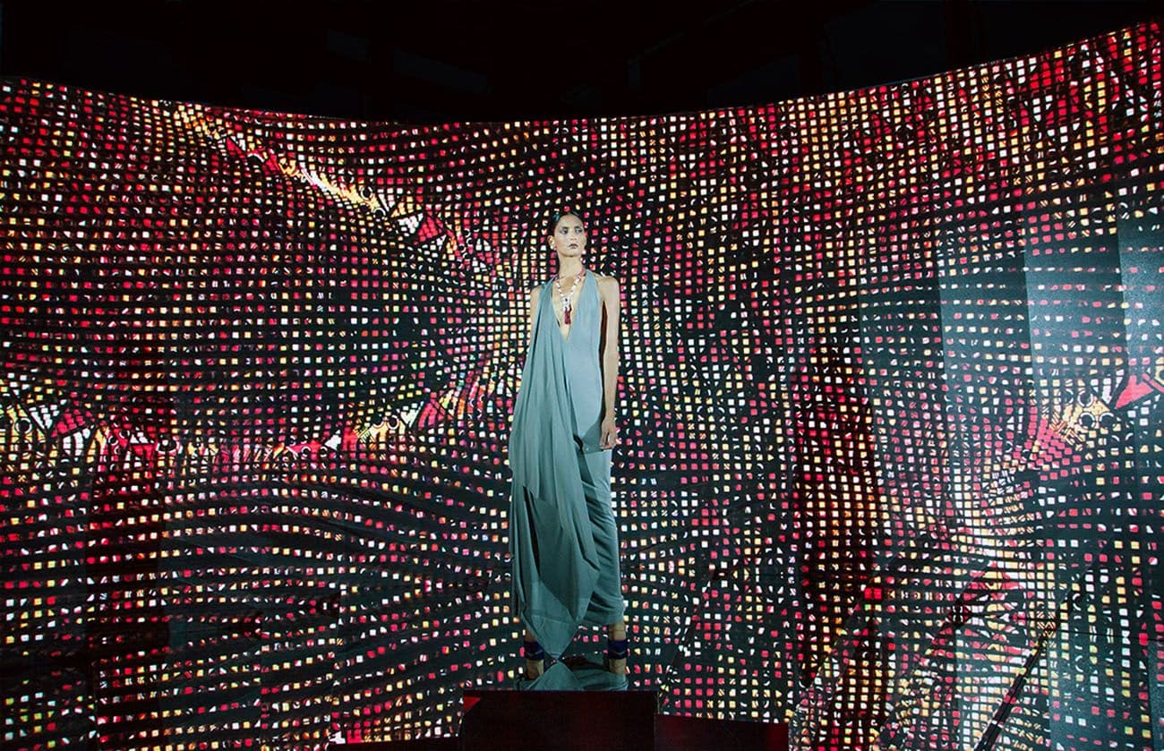 A model in a teal gown standing on a pedestal surrounded by a curved LED screen with a red dotted pattern.