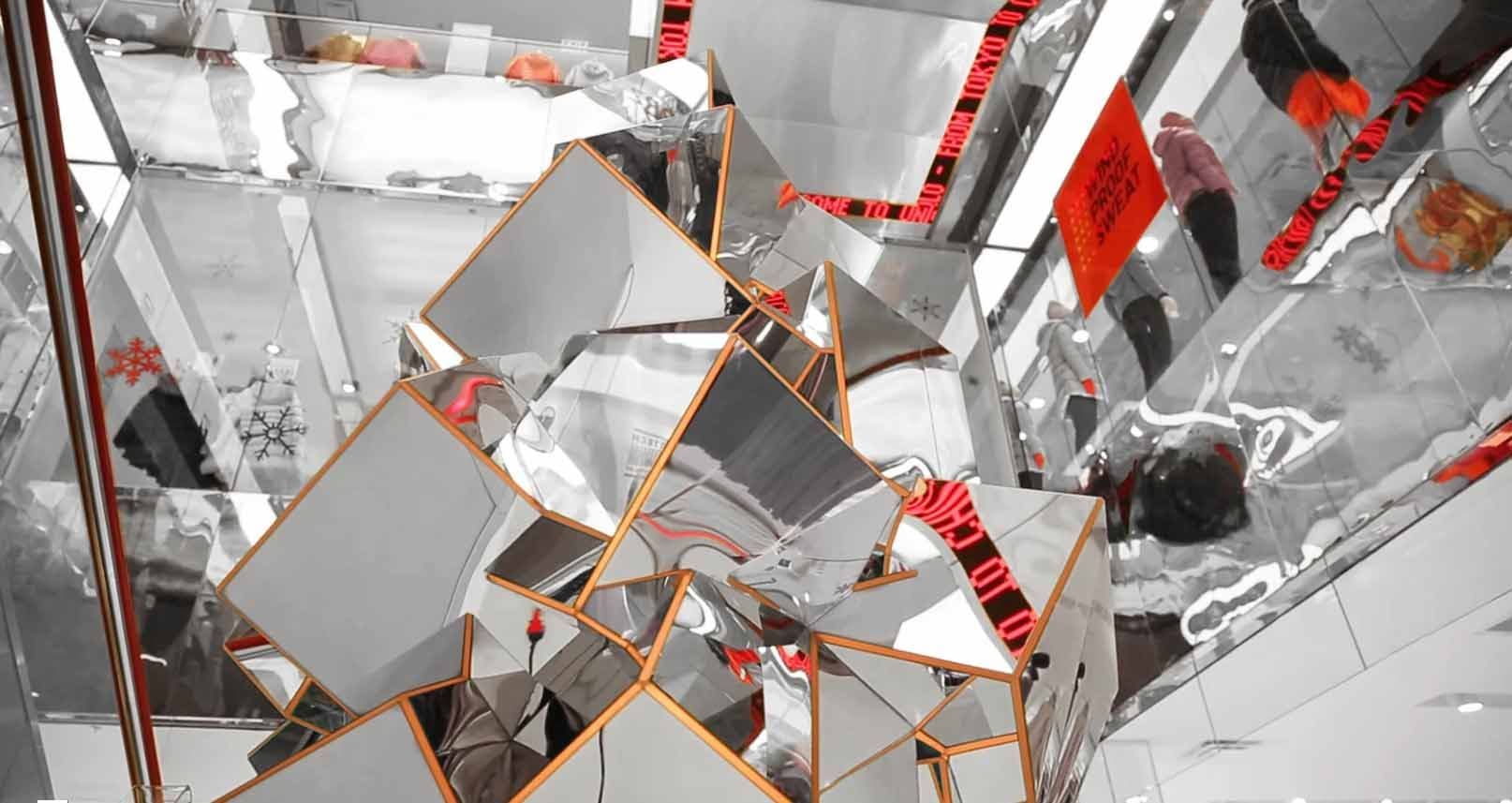 Faceted mirrored sculpture with red-taped edges reflecting the store ceiling and a red-clad figure in the distance.