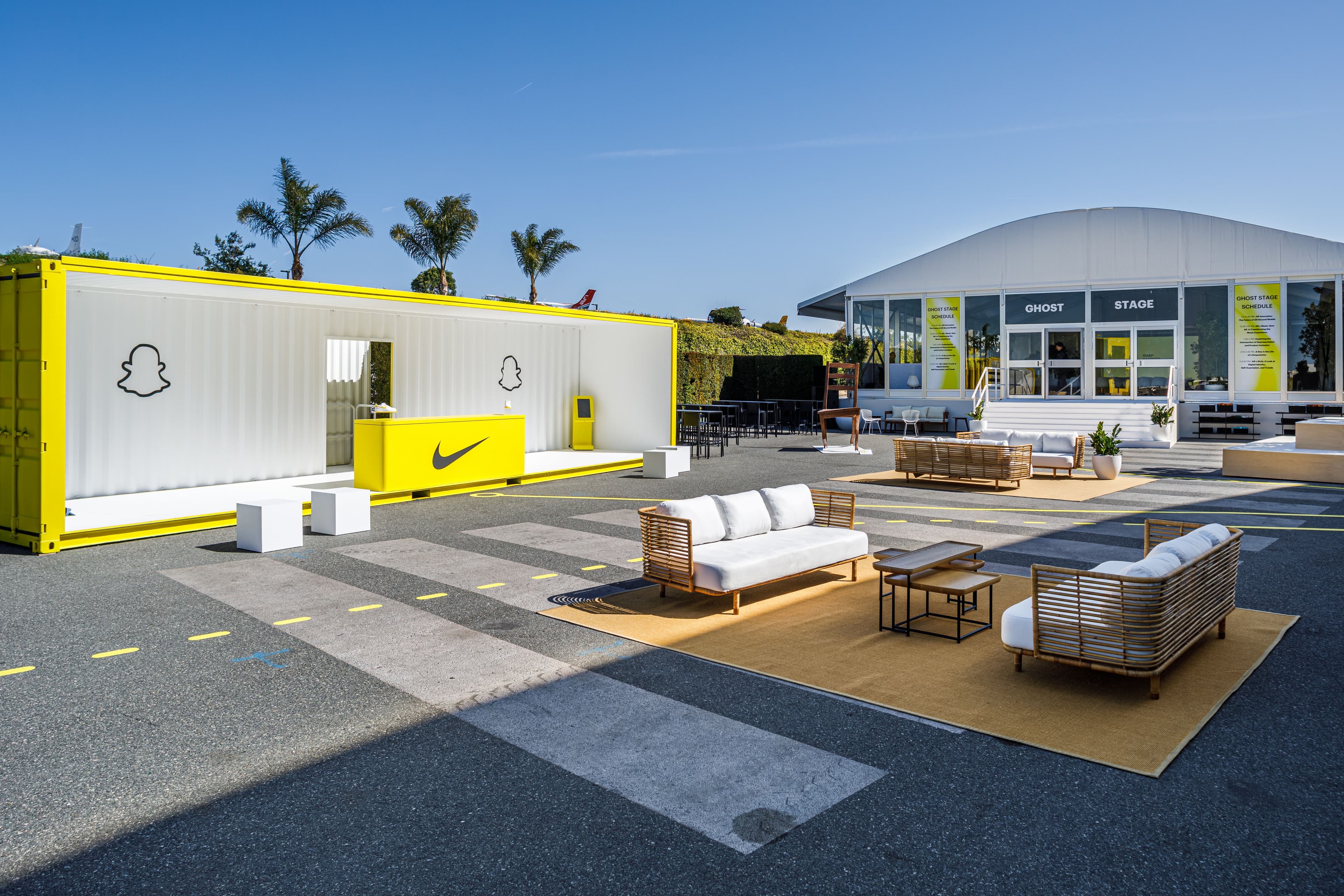 Yellow shipping-container booth with outdoor lounge seating in front, palm trees behind.