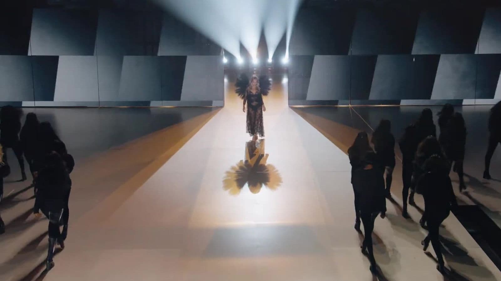 A model in a gold winged costume walks the runway toward the camera between tall angled panel walls, with audience seated on both sides.