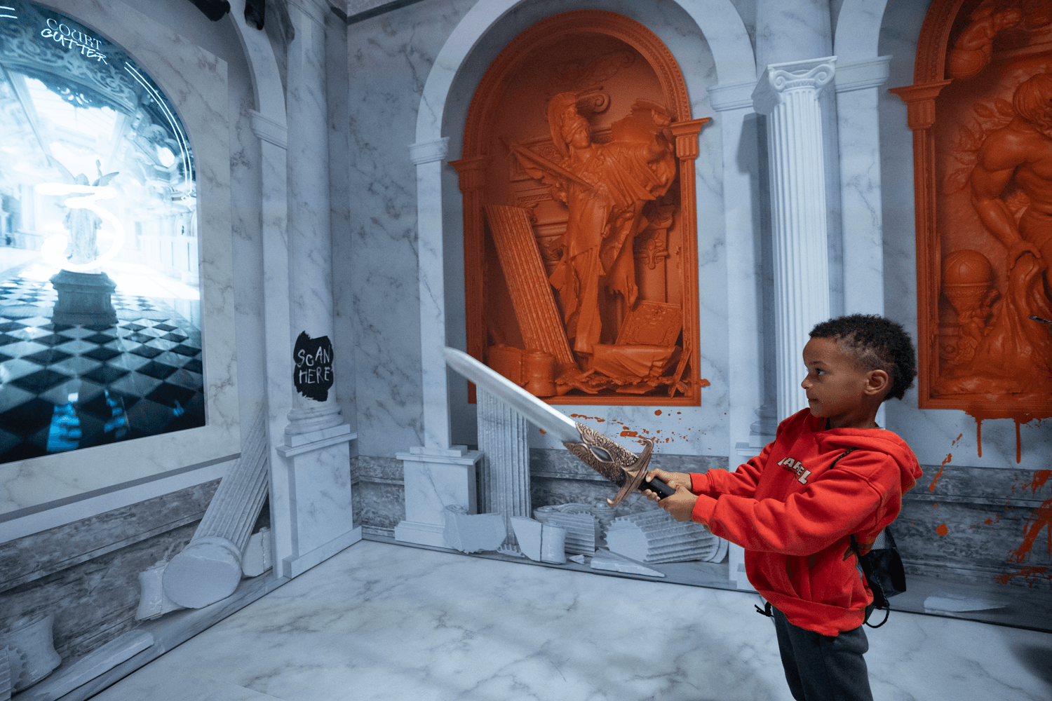 A child in a red hoodie swings a toy sword in a marble hall with orange relief carvings and a checkerboard floor visible in a side alcove.