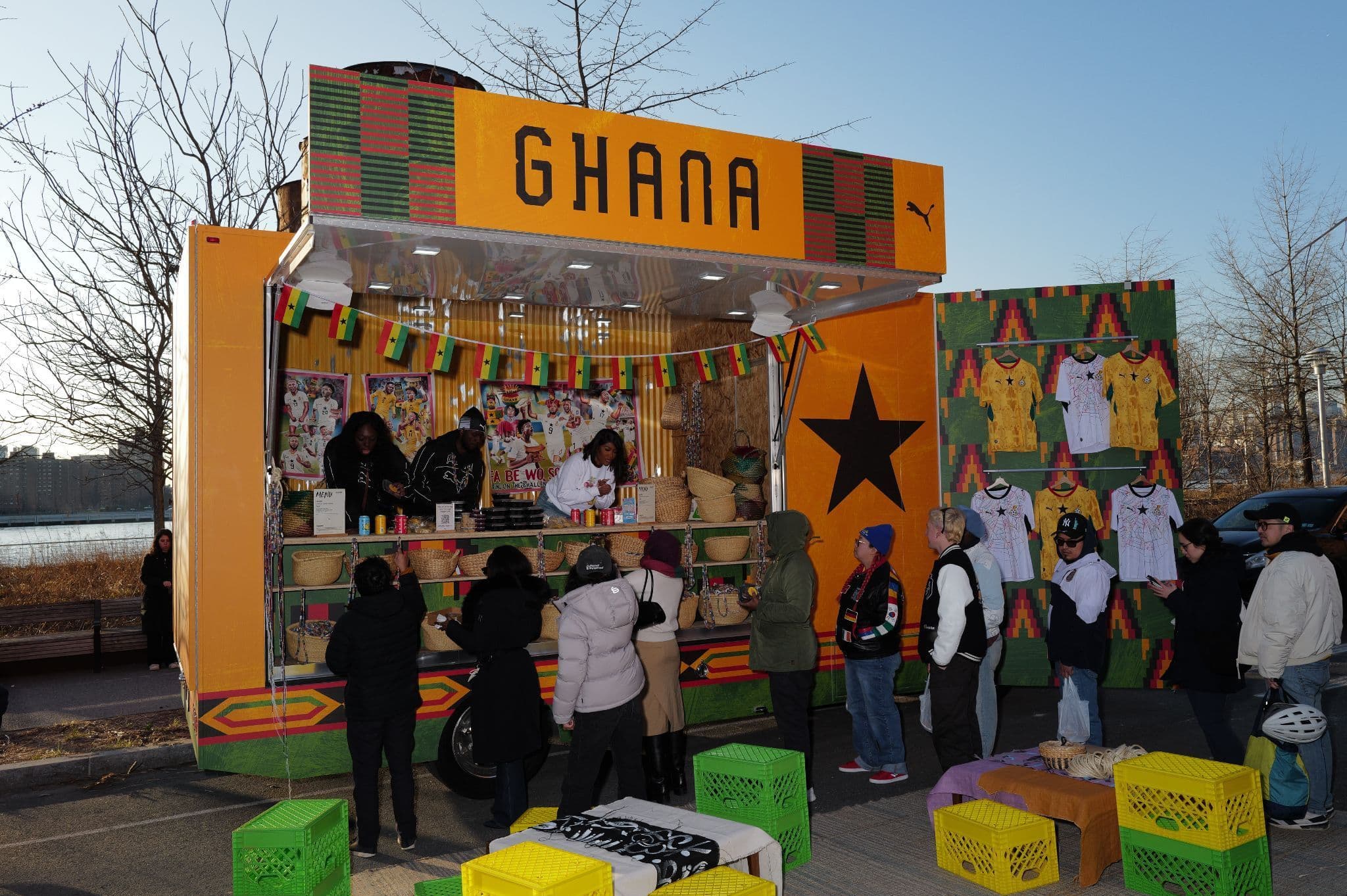 Pop-up shipping-container kiosk branded 'GHANA' with soccer jerseys on display and people gathered around it outdoors.