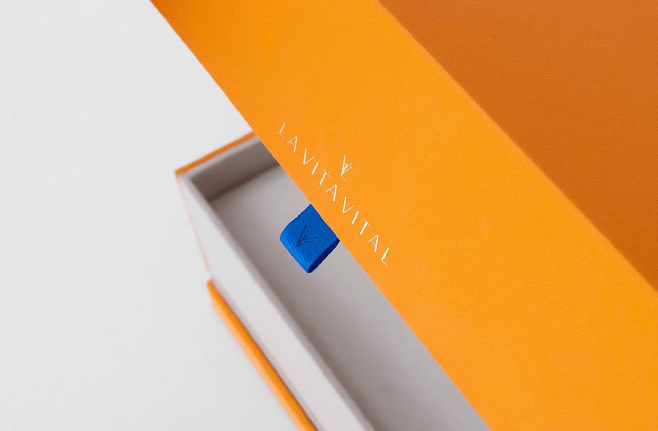 Open orange gift box with a small blue cube nestled inside, next to the La Vita Vital logo embossed on the lid's underside.