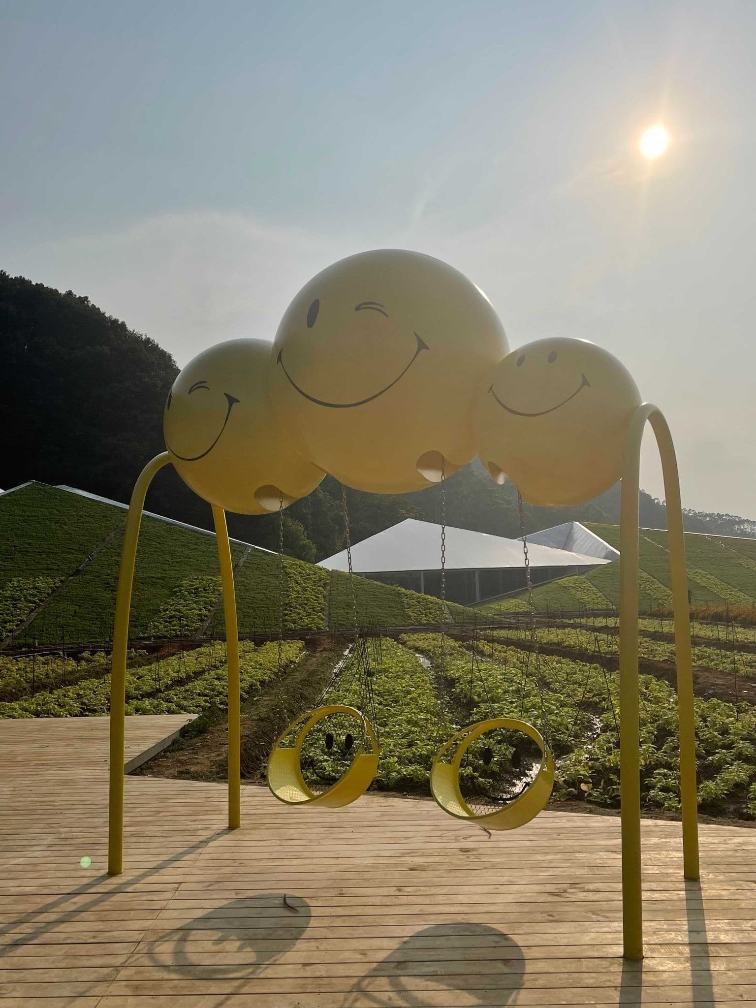 Three connected yellow smiley-face spheres on tall arched posts forming a swing set over a farm field at sunrise.