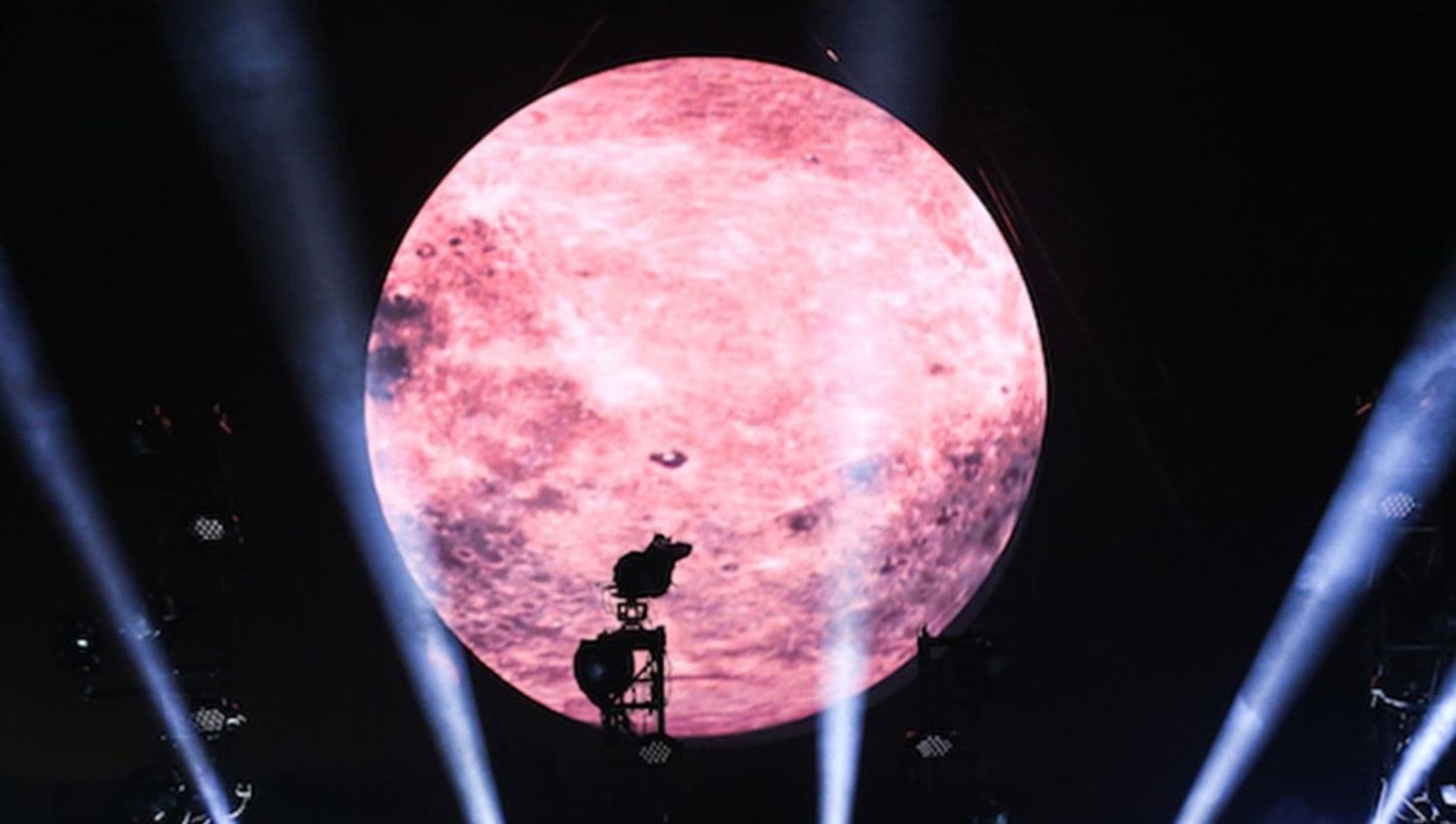 Illuminated pink moon projection above a stage, framed by crossing white spotlight beams at night.