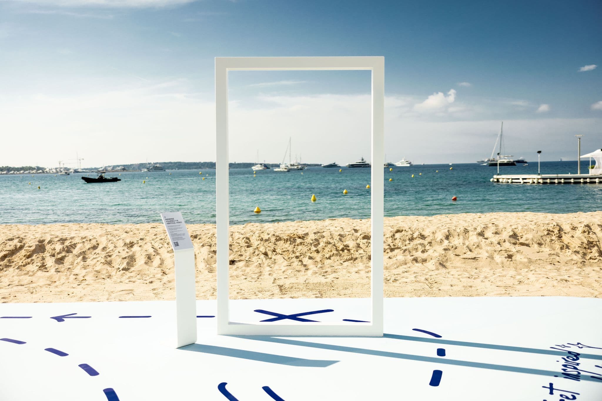 A large white rectangular frame standing on a beach platform with painted blue markings, overlooking sailboats on the water.