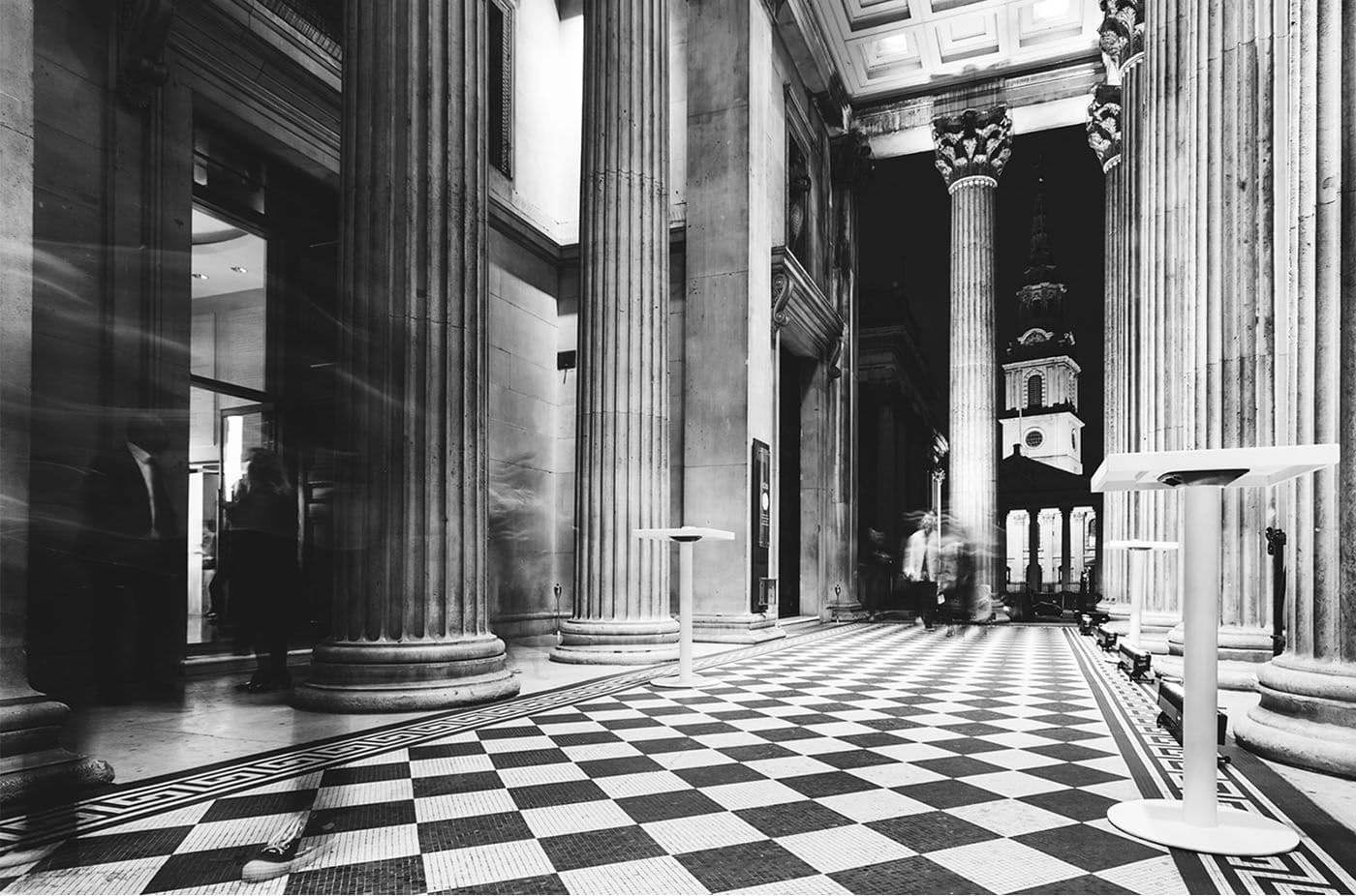 Black-and-white long-exposure photo of a grand colonnaded hall with a checkerboard floor and blurred figures walking.