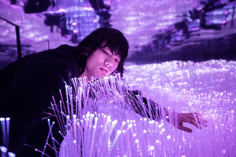 A person walks through a purple fiber-optic light installation, reaching out to touch the hanging strands.