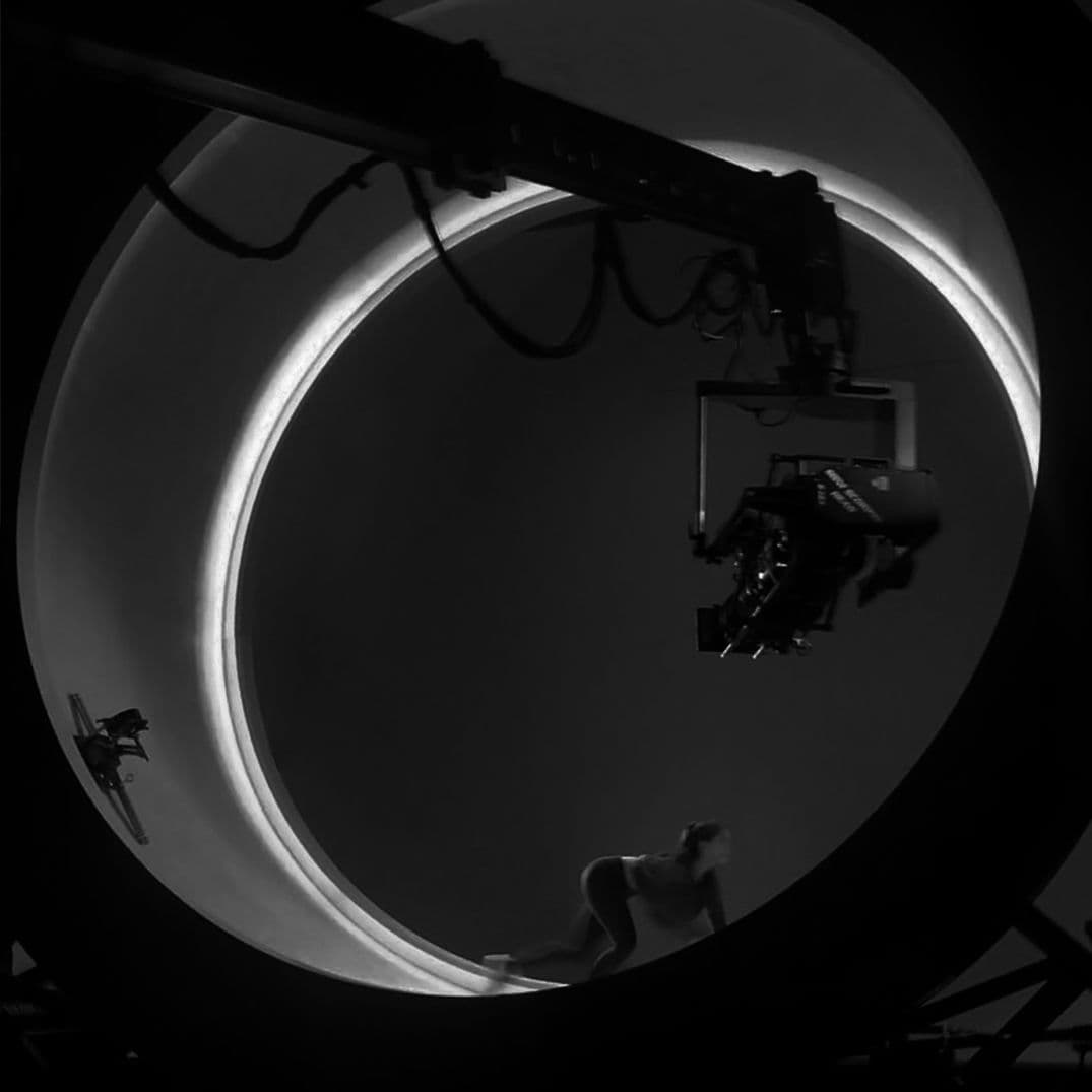 Black-and-white behind-the-scenes photo of a film crew operating a camera crane inside the circular ring set, with two performers visible below.