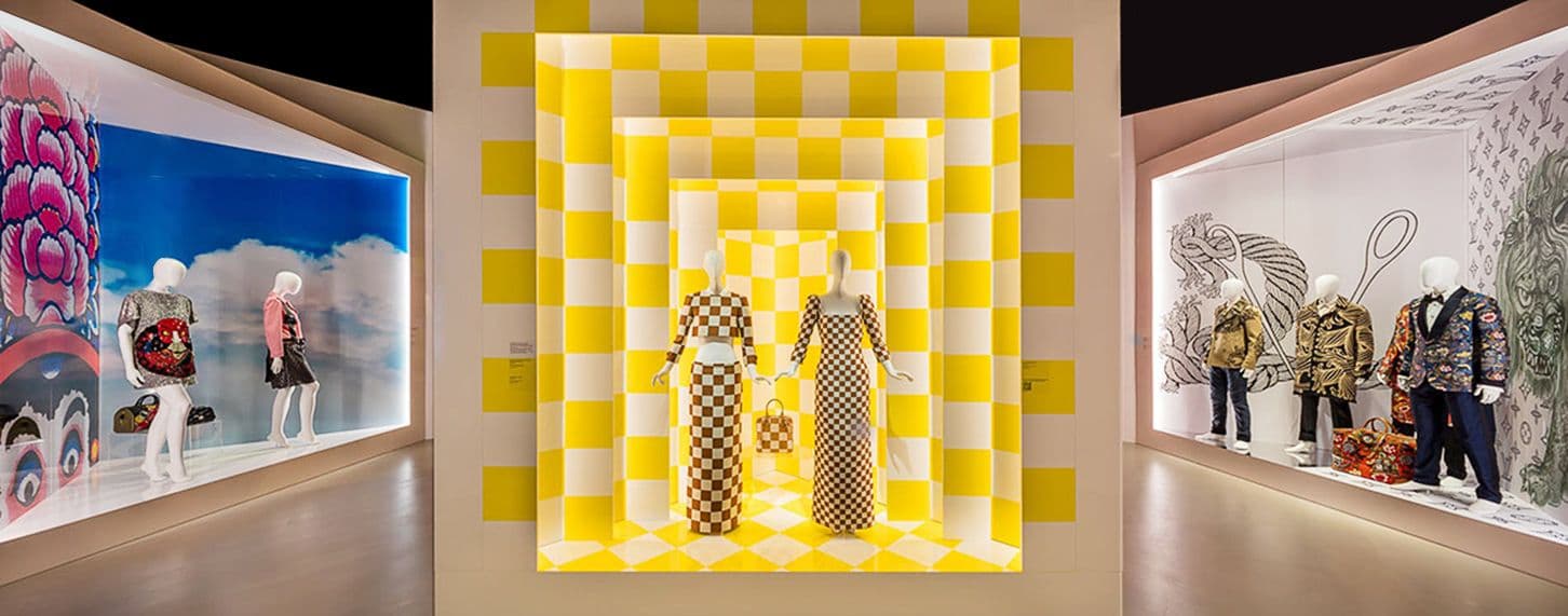 Illuminated yellow checkerboard display case flanked by mannequins in colorful outfits behind glass storefront windows.