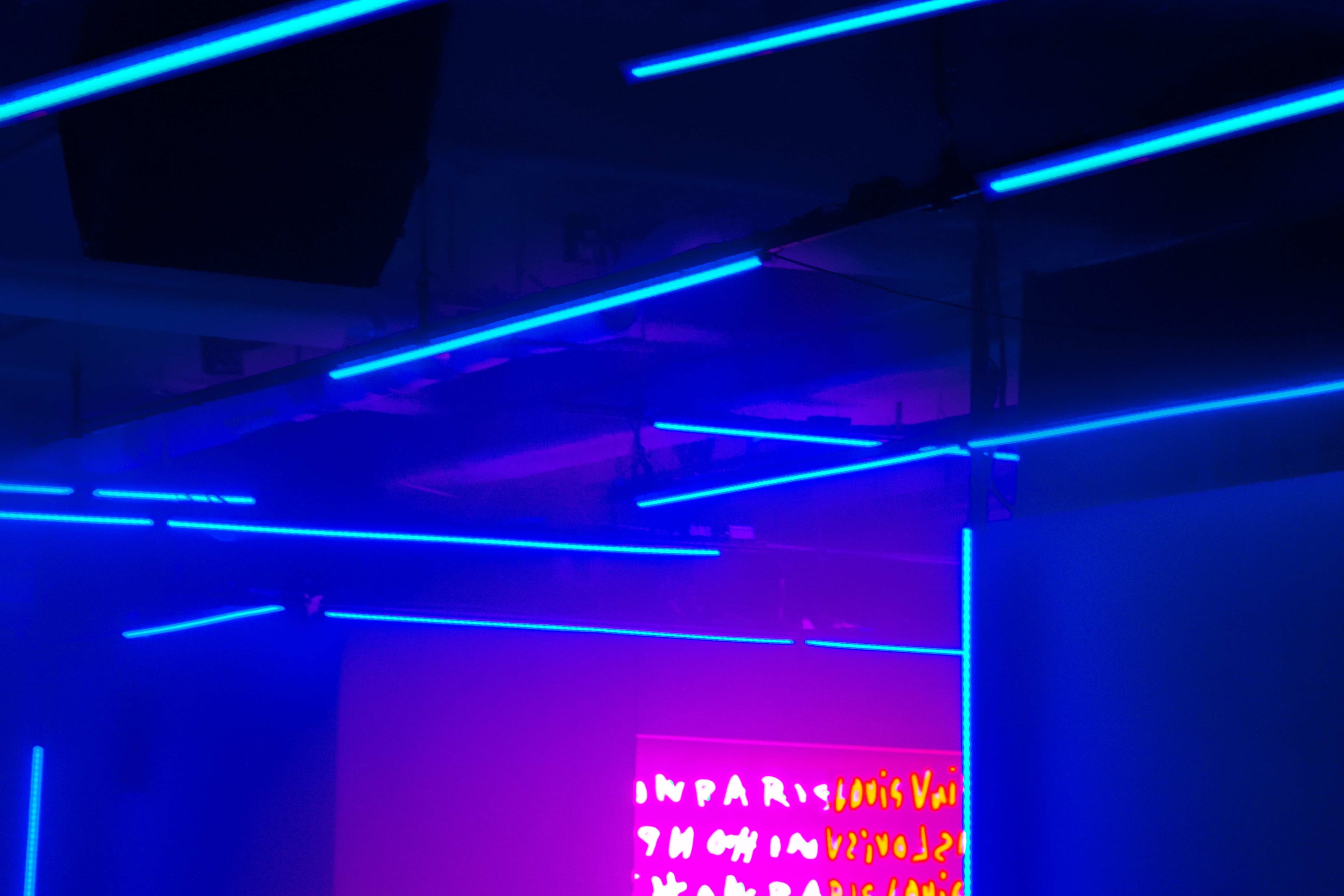 Blue neon tube installation crossing a ceiling above a glowing pink and orange text display on the wall.