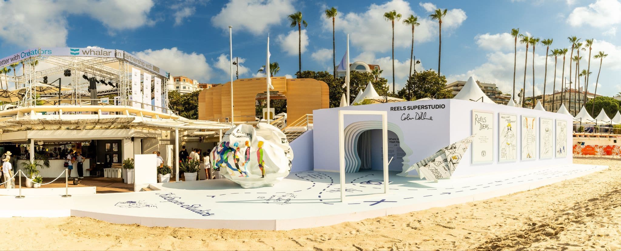 An outdoor beach installation with a tunnel entrance and a large sculptural white ball, palm trees and tents in the background.