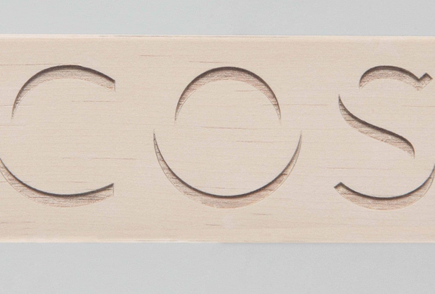 Close-up of the COS wordmark cut into a pale wood plank.