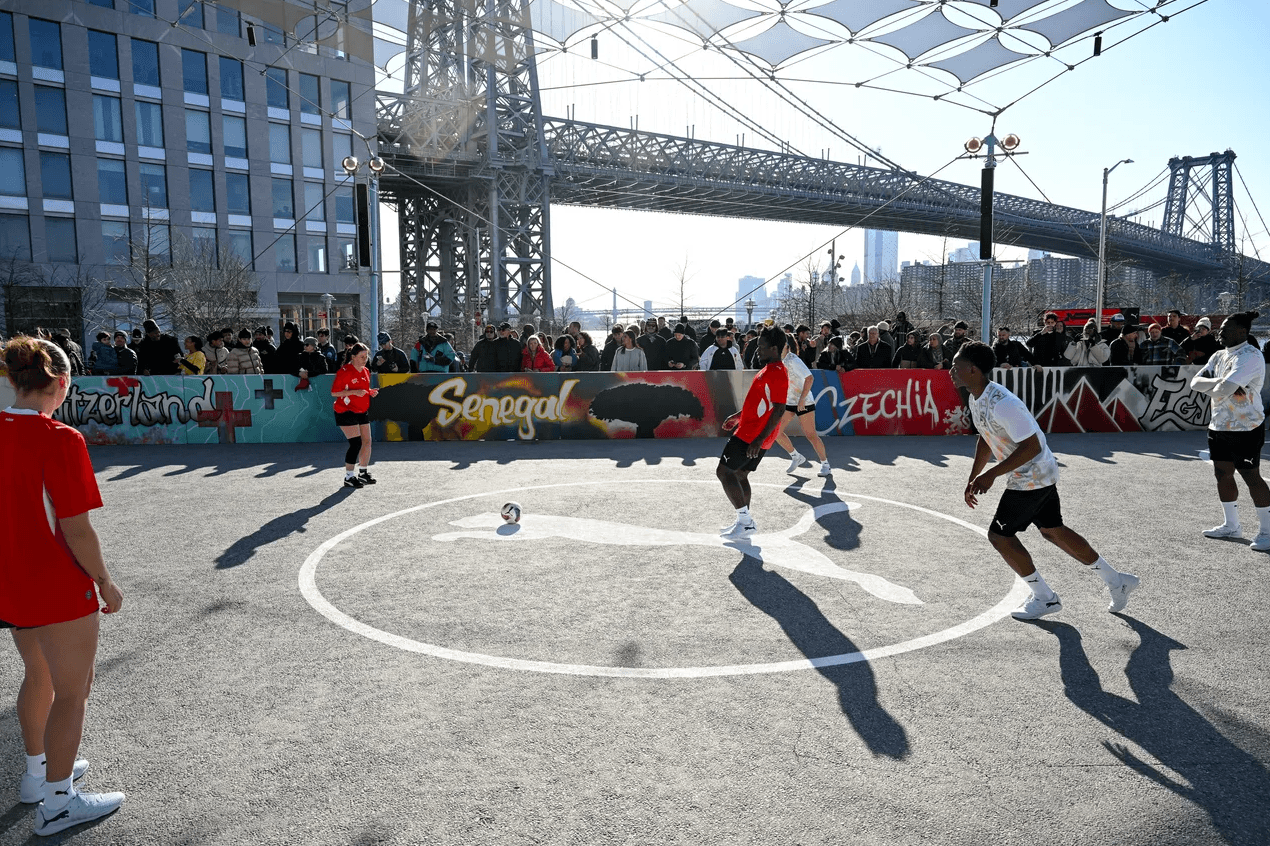 Two teams playing basketball on an outdoor court beneath a suspension bridge, watched by a small crowd along the sideline.