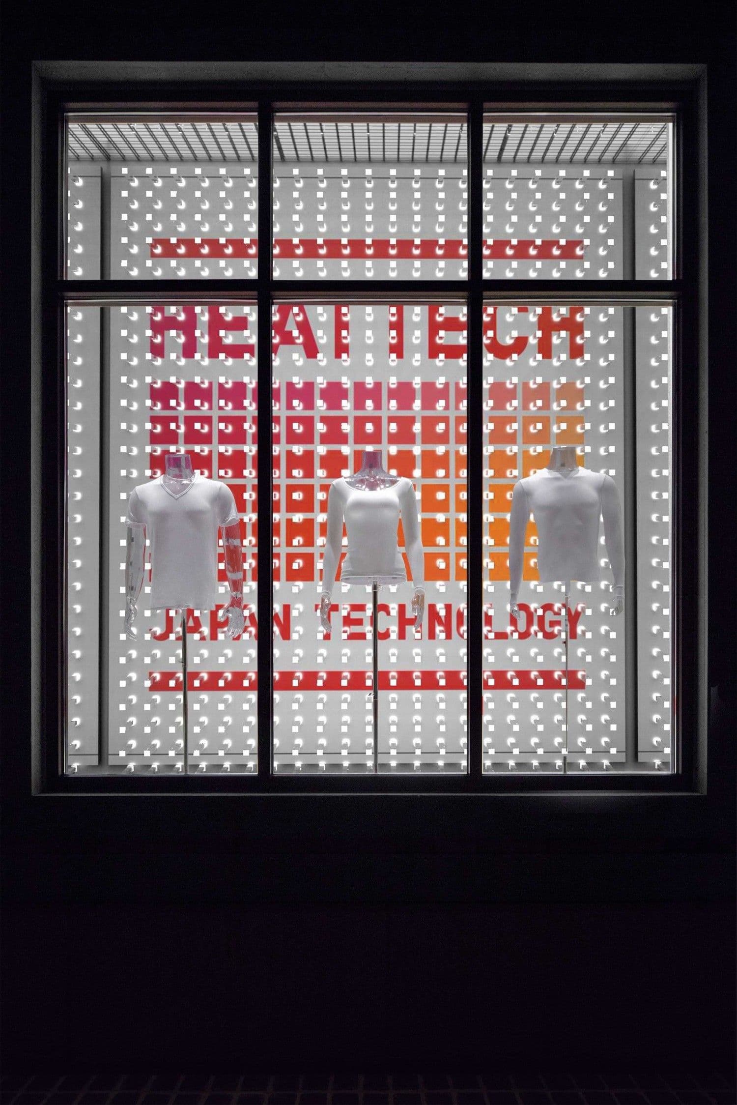 Dark storefront window lit with a perforated dot-matrix pattern spelling HEAT TECH and Japan Technology above three lit mannequin torsos.
