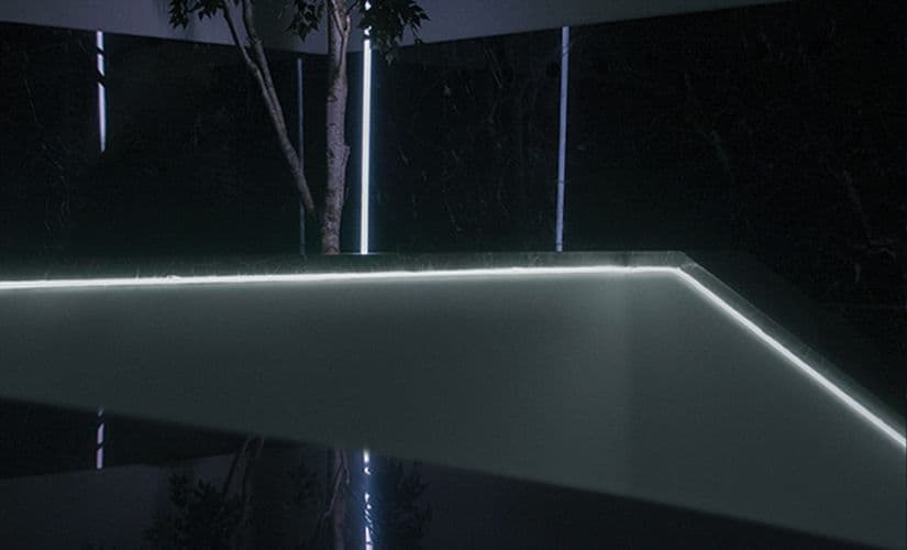 Night shot of a glowing white plinth edge reflected on a glossy floor, with a lit tree in the background.