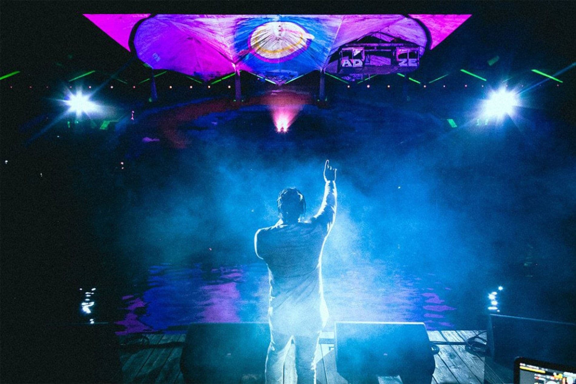 Silhouette of a person raising an arm on a fog-filled stage beneath a colorful circular canopy projection at night.