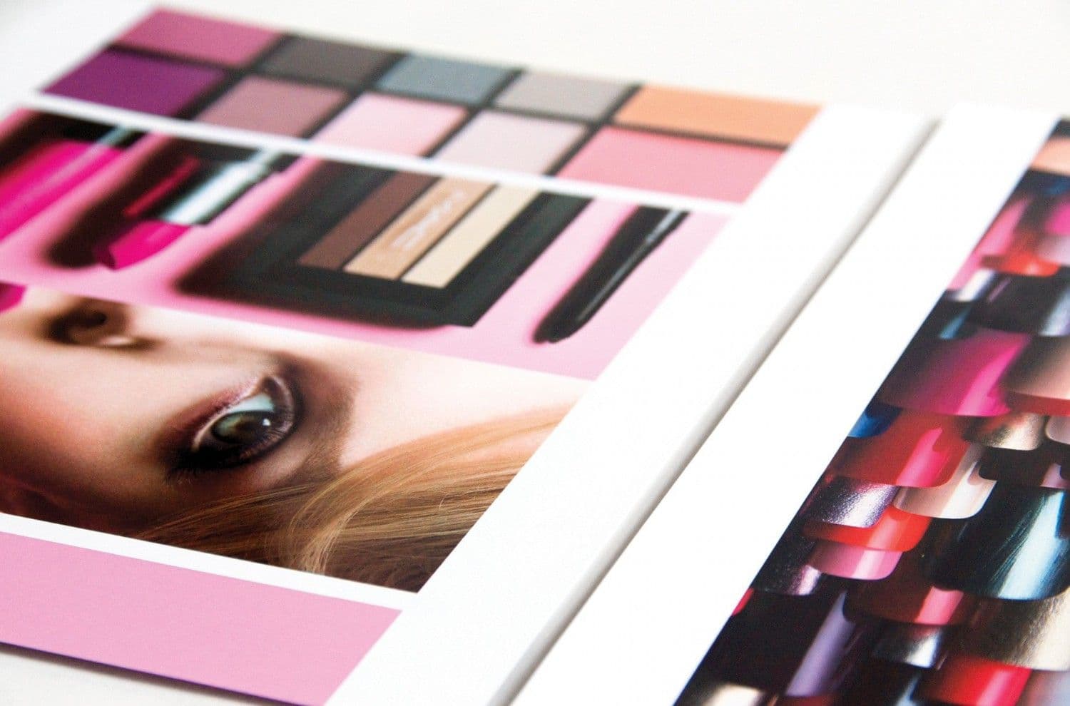 Fanned stack of catalog pages showing eyeshadow palettes, a close-up model eye, and rows of lipstick swatches in pink and red tones.