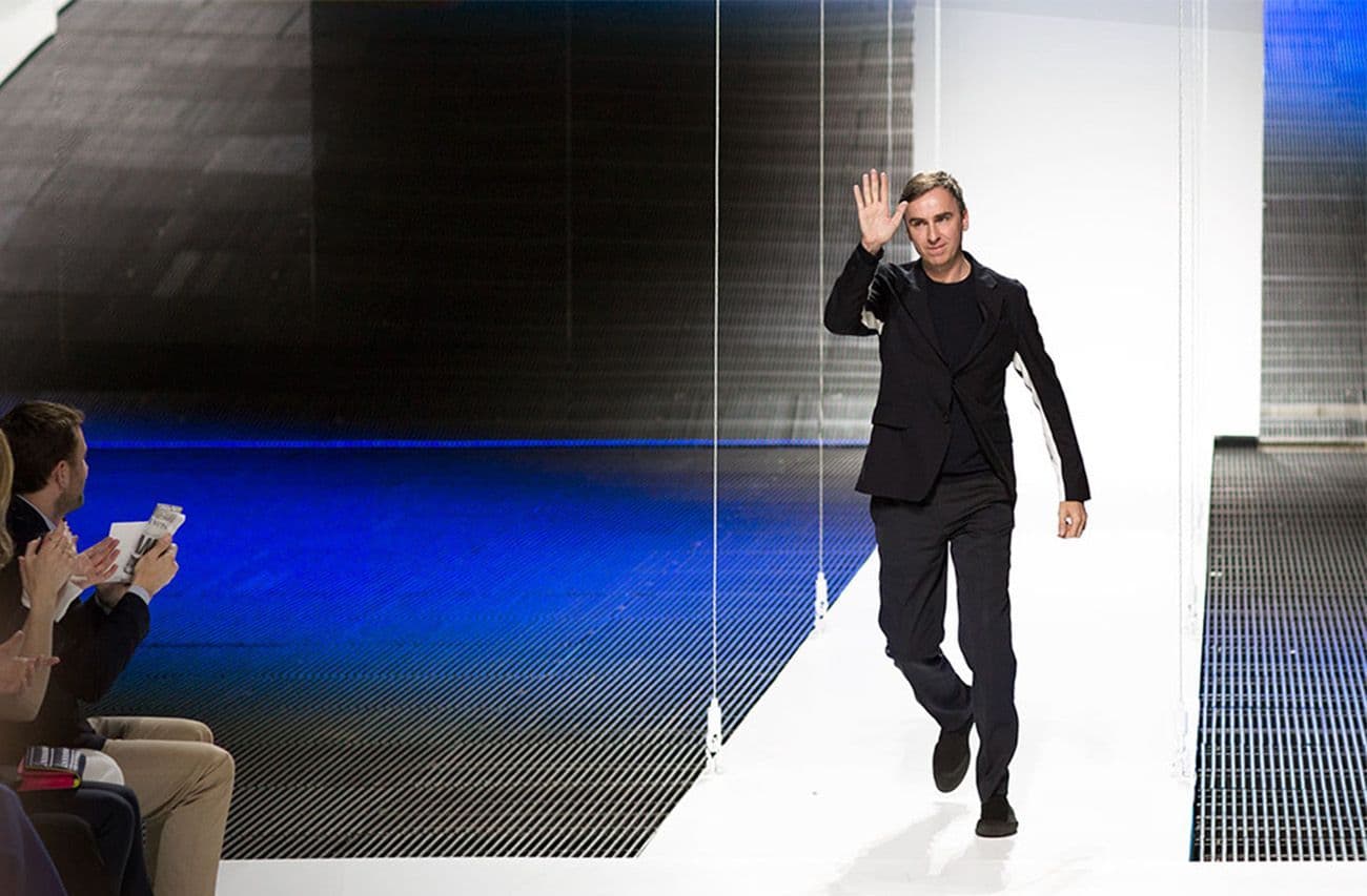 A man in an all-black outfit walks the runway, waving to the audience, against a dark blue backdrop.