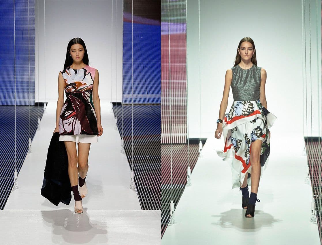 Two models walk together, one in a graphic floral-print dress and the other in a pleated grey skirt, against a dark backdrop.