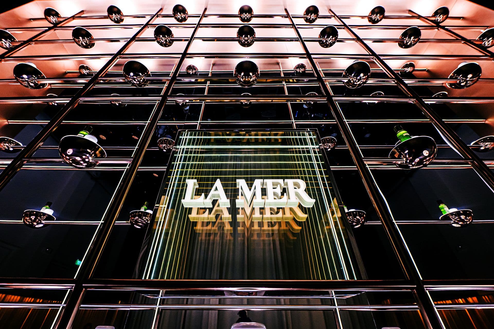 Rows of glass bottles suspended in a grid overhead, with an illuminated La Mer sign at the center, viewed from below.