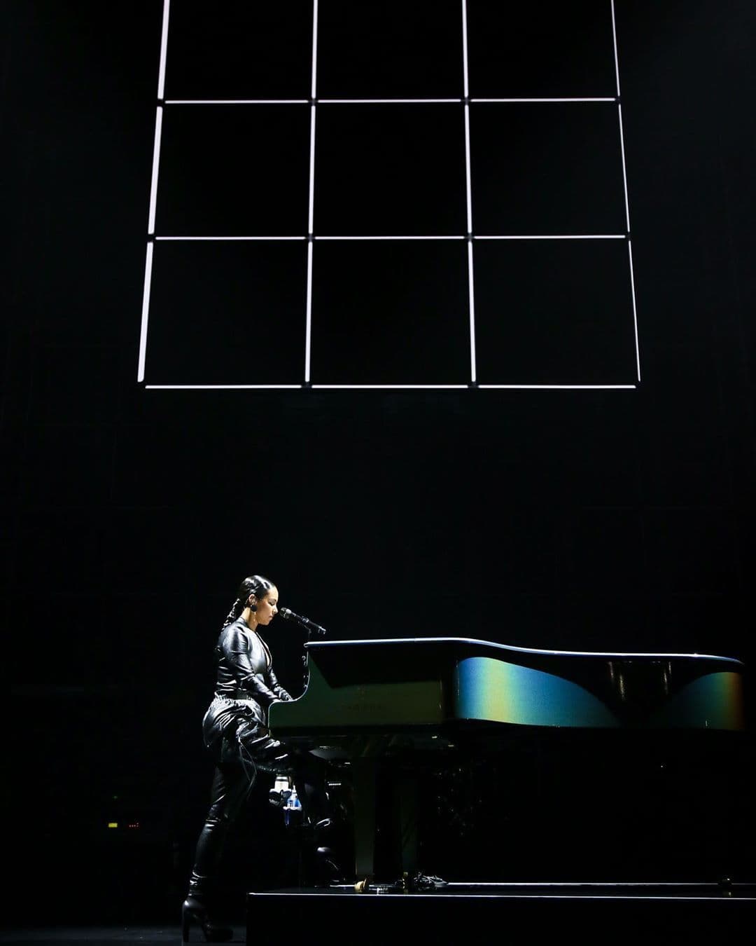 Performer singing at a grand piano on a dark stage lit from below, with the wireframe grid structure visible overhead.