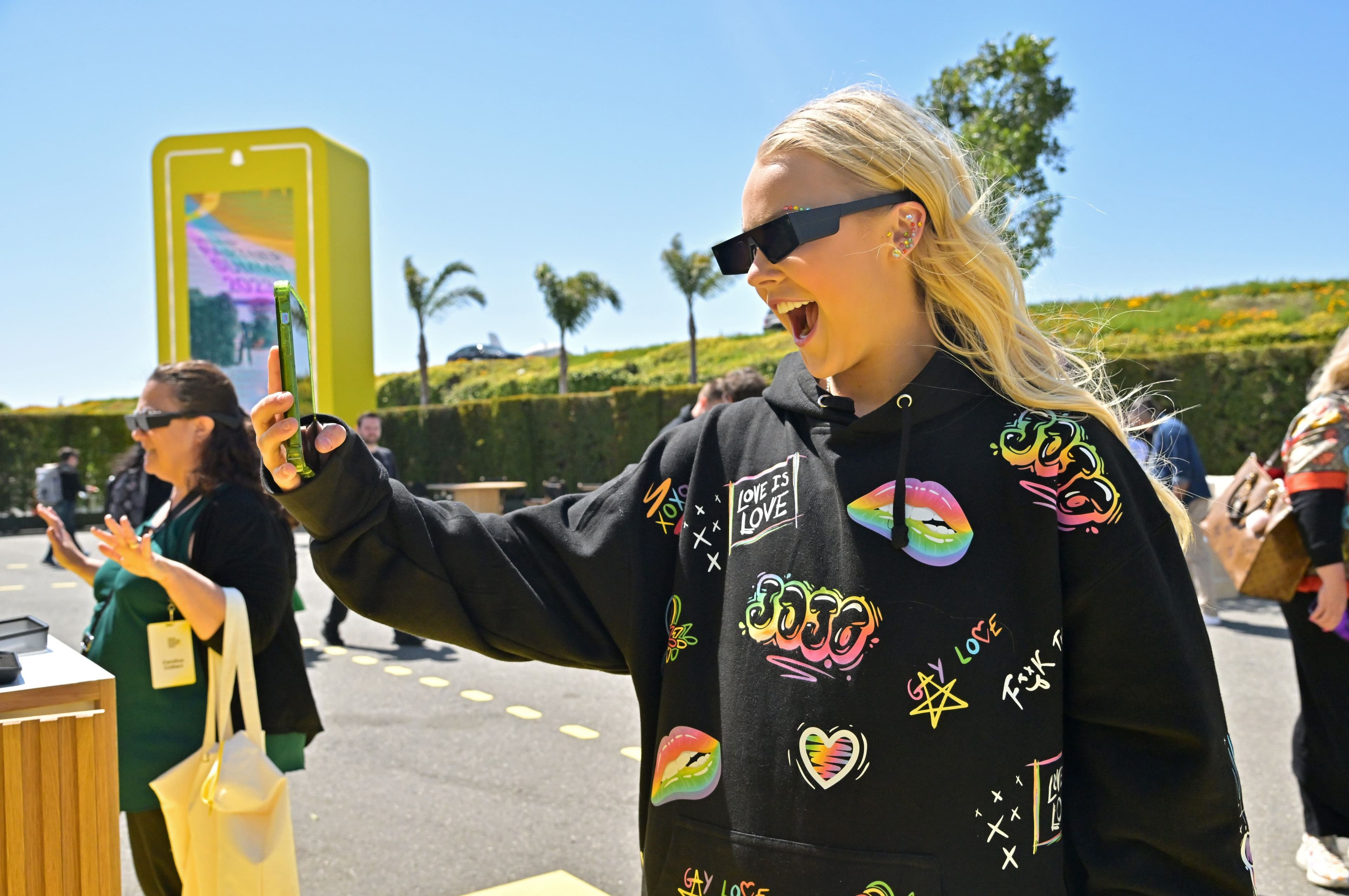 Woman wearing patterned jacket and smart glasses taking a photo with her phone outdoors.